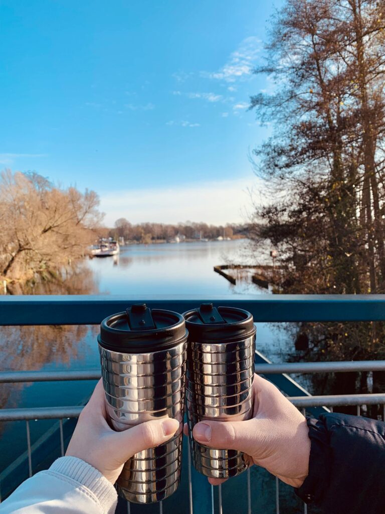 Two people holding reusable mugs by a serene lake on a sunny day.