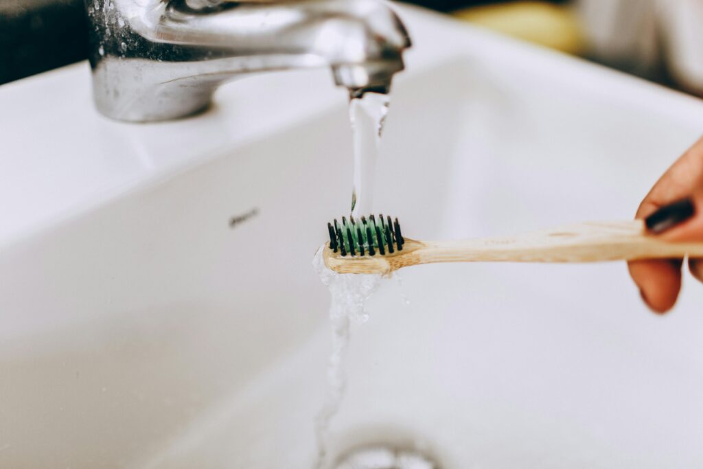 Eco-friendly bamboo toothbrush under running water from faucet in a bathroom sink.