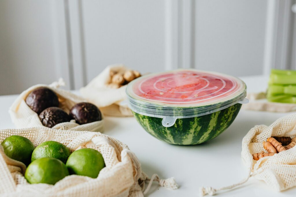 pexels-photo-5843555-5843555 A fresh sliced watermelon with reusable food wrap, surrounded by avocados and limes on a white table.