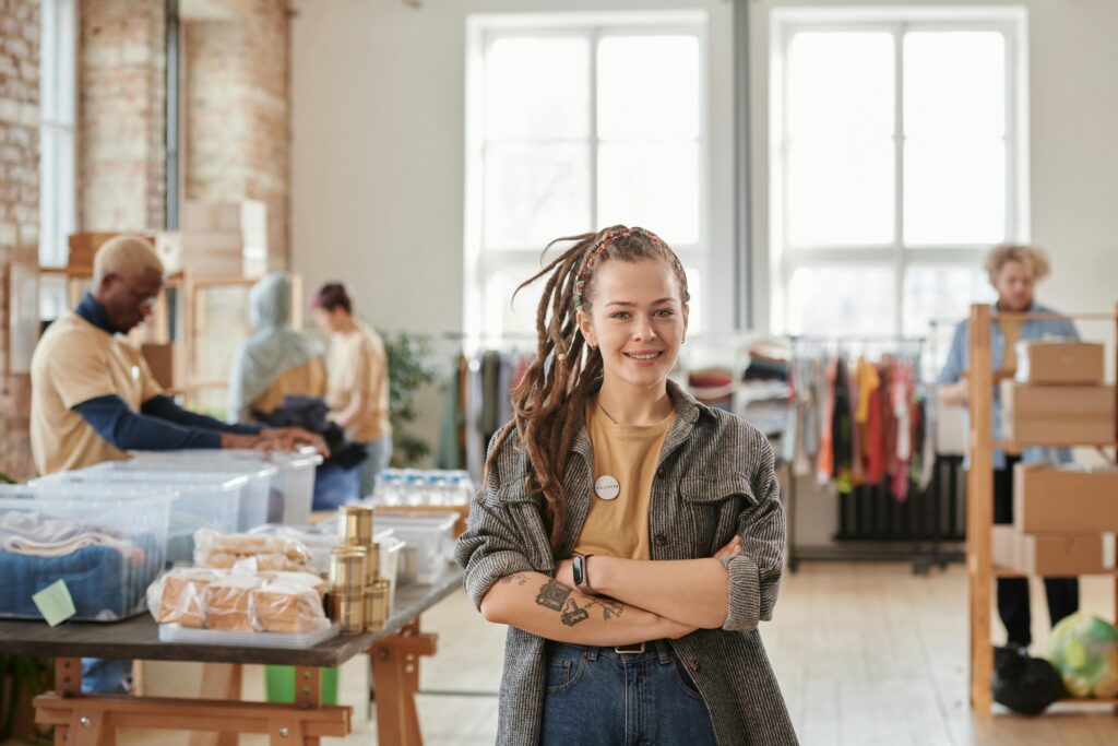 Smiling woman with dreadlocks standing confidently in a thrift store, surrounded by busy attendants.