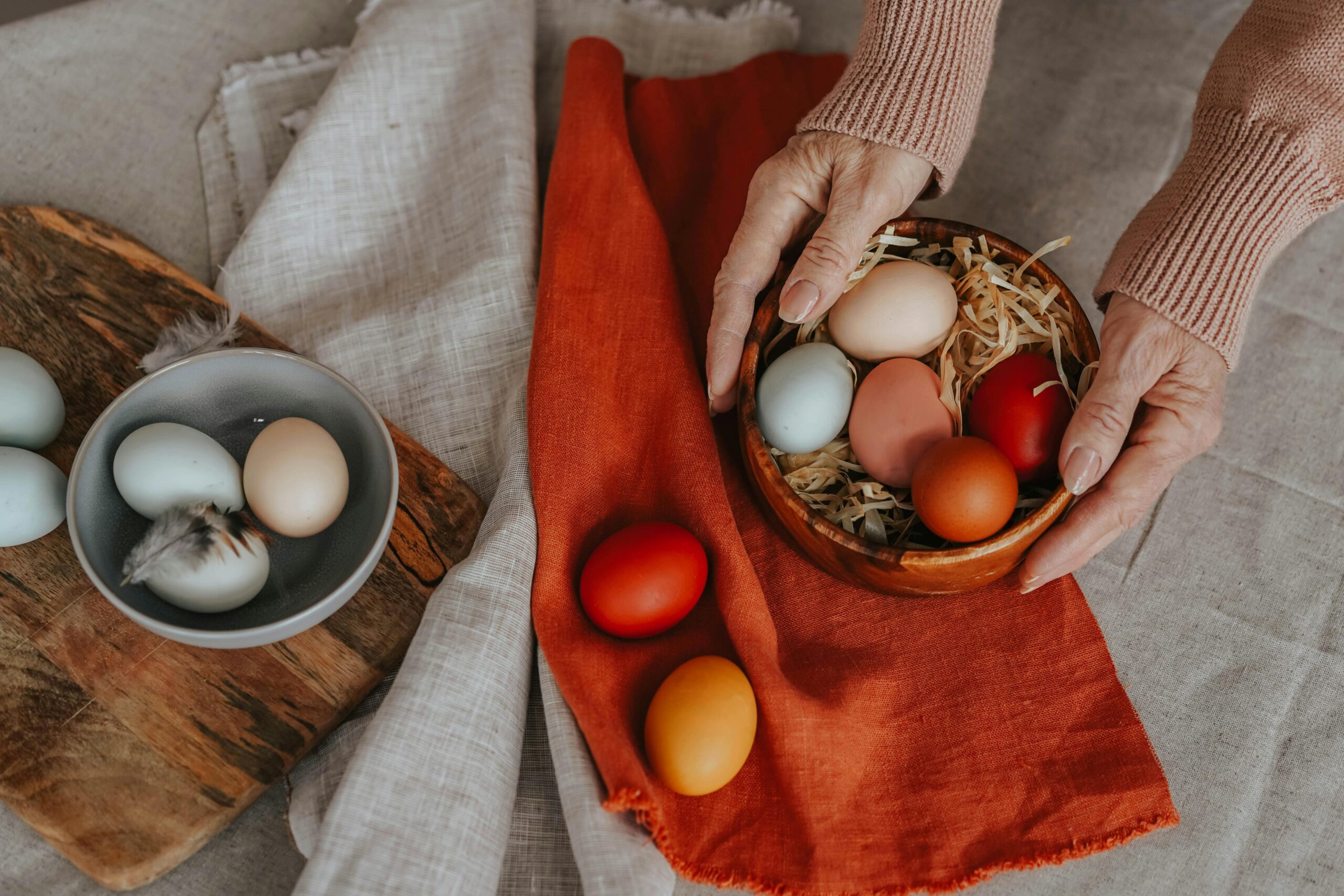 Hands arranging colorful Easter eggs in wooden bowls on a rustic table setup.