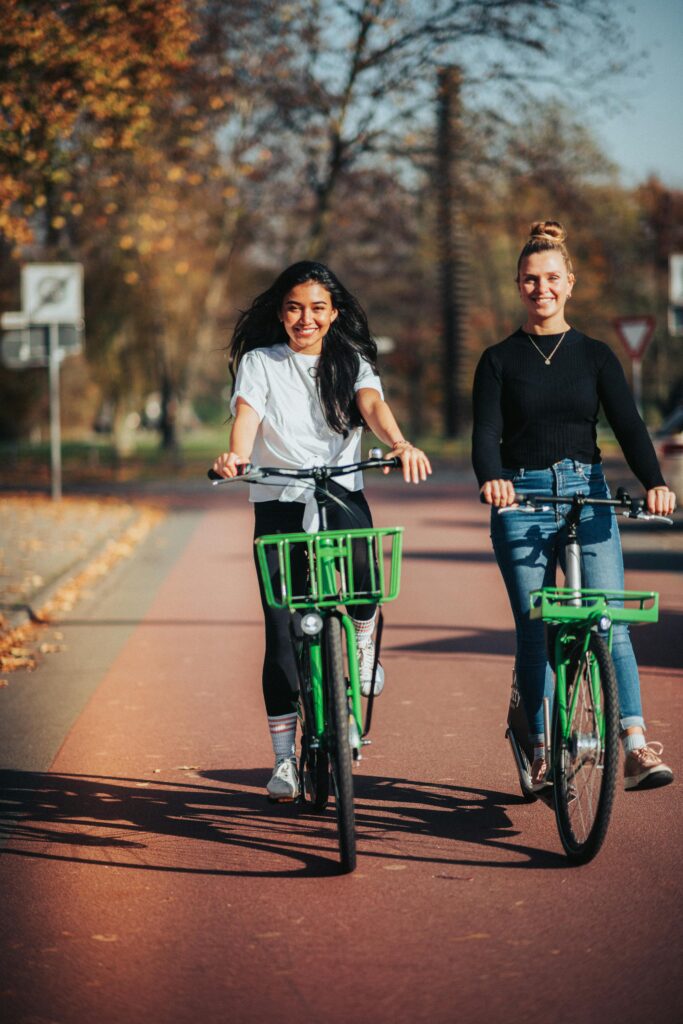 Two women riding green bicycles on a sunny day in a park.